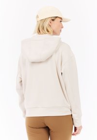 Lichtbeige hoodie met een trekkoordkap, voorzien van een relaxte pasvorm en zichtbare stikseldetails. Gecombineerd met bruine leggings.