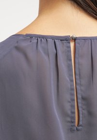 Blusa grigia con chiusura a apertura a chiave sul retro, arricchita da un bottone decorativo. Tessuto morbido e strutturato con dettagli arricciati sul collo.
