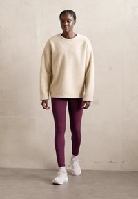 Maglione oversize in crema e texturizzato abbinato a legging bordeaux aderenti e scarpe da ginnastica bianche. Sullo sfondo c'è un tono neutro chiaro.