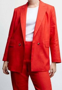 Una persona indossa un giubbino rosso brillante e pantaloni coordinati su una camicia bianca, mostrando un outfit elegante e moderno su uno sfondo neutro.