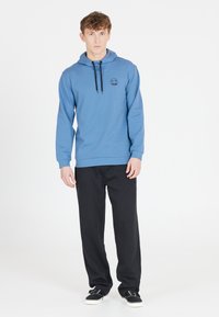 Ljusblå huvtröja med en logo, dragsko vid halsen och ribbad nederkant. Kombinerad med svarta byxor och svarta skor, stående mot en vit bakgrund.