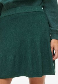 Fustă tricotată de culoare verde închis, cu un talie cu nervuri. Designul evazat prezintă o textură moale și țesătură ușor plisată la margine.