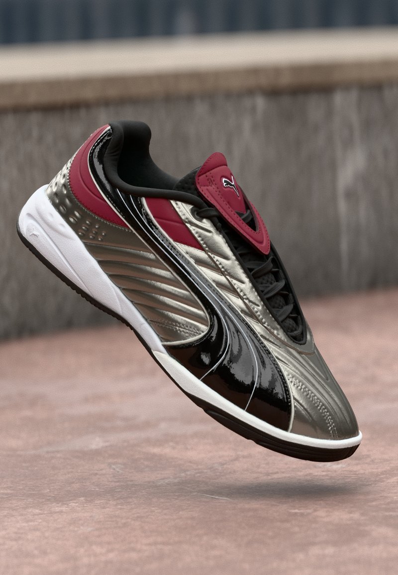 Baskets Puma métalliques argentées, noires et rouges flottant au-dessus d'un sol rosé avec un mur flou en arrière-plan.