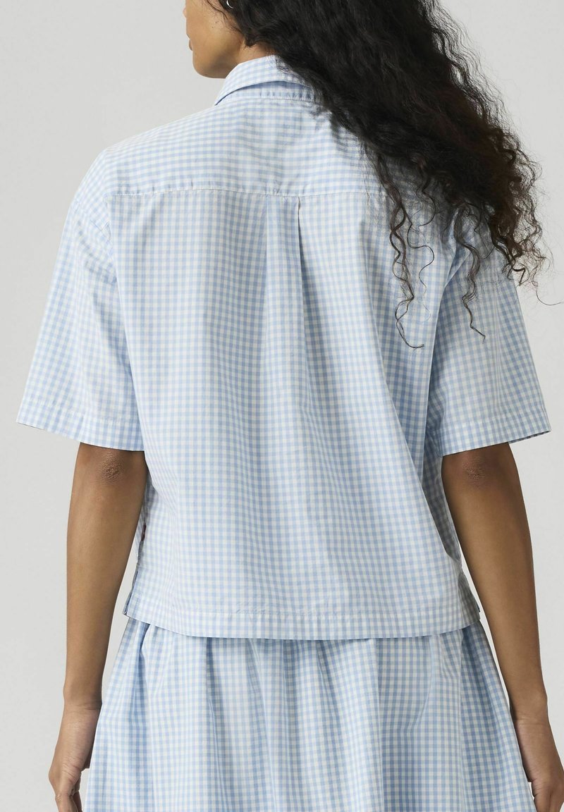 Femme aux longs cheveux bouclés portant une chemise à manches courtes vichy bleu clair et blanc et une jupe assortie, vue de dos.