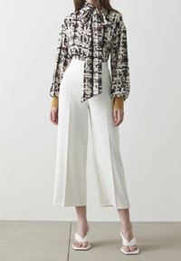Mønstret bluse i sort og hvid med krave og sløjfe, parret med brede hvide bukser og hvide sandaler med et snoet design.
