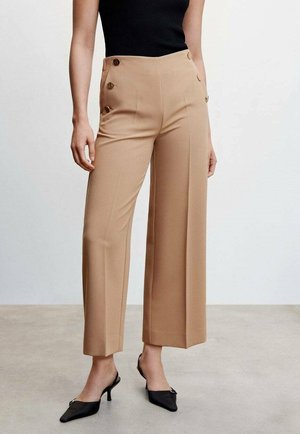 Kobieta ubrana w beżowe szerokie spodnie z wysokim stanem i guzikami po bokach, czarny bezrękawnik oraz czarne szpilki z ostrymi noskami, stojąca na szarej podłodze.