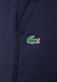 Tummansininen kangas, jossa on sileä rakenne, vasemmalla puolella vihreä ja punainen krokotiililogo. Muotoilu sisältää sivutaskun.