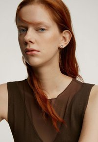 Top marrón sin mangas con un diseño acanalado texturizado. La modelo tiene el cabello rojo de longitud media y lleva un simple aro de arete redondo.