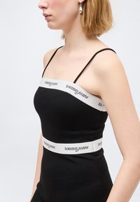 Γυναίκα με μαύρο αμάνικο τοπ με λεπτές τιράντες και λευκές ταινίες με το λογότυπο "tommy jeans" γύρω από το στήθος και τη μέση, ανοιχτό φόντο.