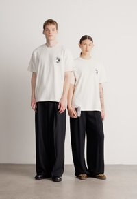 Bele, prevelike majice s potiskanim logotipom spredaj, kombinirane z ohlapnimi črnimi hlačami. Eden drži kovinski torbico; oba nosita priložnostne čevlje.