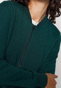Prošívaná zelená bomber bunda s kulatým výstřihem, předním zipovým zapínáním a žebrovanými manžetami. Texturovaná látka s diamantovým vzorem.