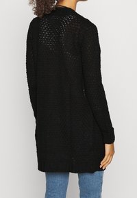 Žena v dlouhém černém otevřeném pleteném kardiganu přes světle modré džíny, zobrazená zezadu na jednobarevném pozadí.