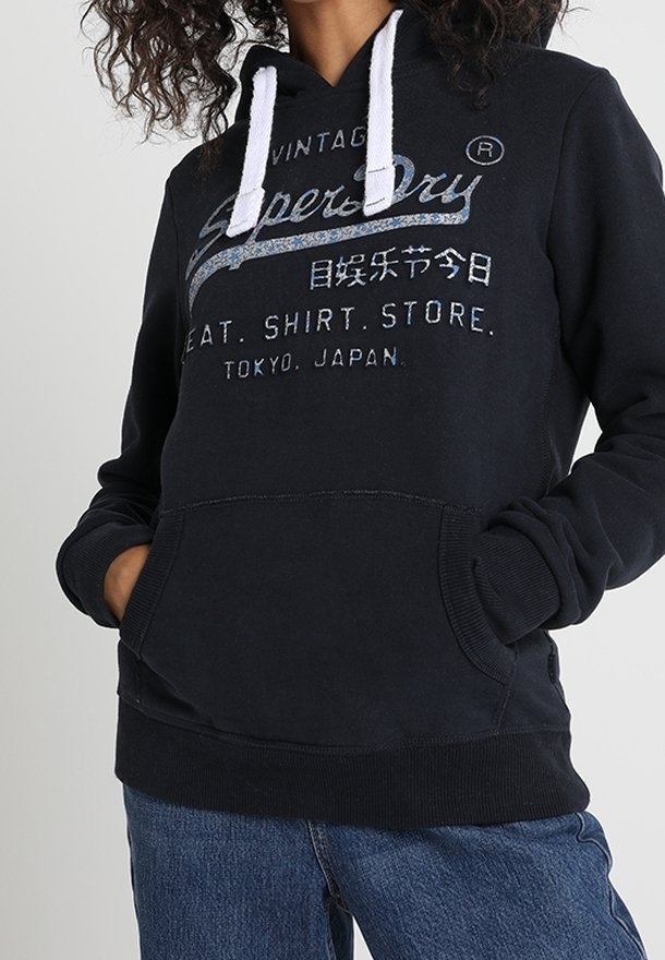 Sudadera negra con cordones blancos, con un bolsillo frontal, tela texturizada y logo azul impreso. Combinada con jeans de mezclilla.