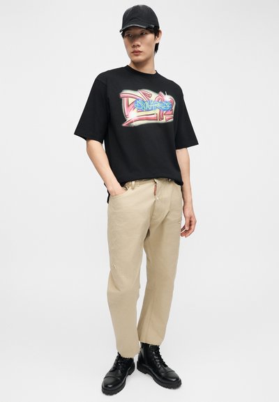 Jeune homme portant une casquette noire, un t-shirt graphique style graffiti noir, un pantalon beige coupe décontractée et des bottes noires à lacets, debout avec une main dans la poche.