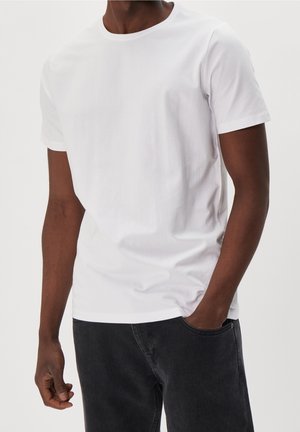 Homme portant un T-shirt blanc uni à manches courtes et un jean noir, avec une main dans la poche, sur fond blanc.