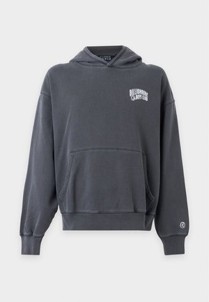 Młody mężczyzna w czarnej czapce Billionaire Boys Club, ciemnoszarej bluzie z kapturem na koszulę w kratę, czarnych dżinsach i czarnych butach, stojący na białym tle.