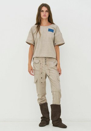 Beige linnen top met een blauw zakje, gecombineerd met beige cargobroek met meerdere zakken en een trekkoord in de taille, gedragen met donkerbruine laarzen.