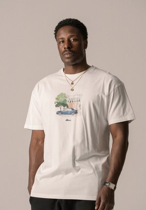 White cotton t-shirt featuring a detailed graphic of a blue car and green tree, with a small text logo beneath the illustration.