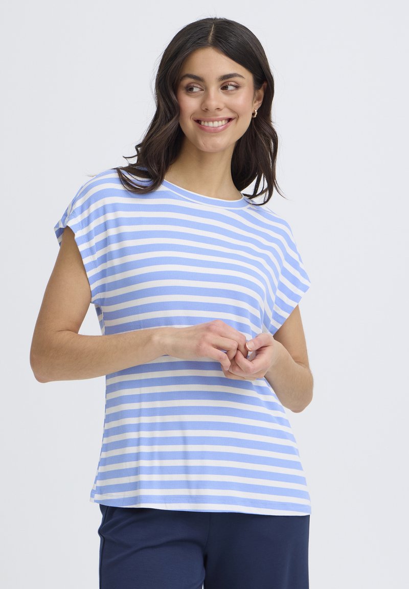 Femme souriante, portant une chemise à manches courtes rayée bleu clair et blanc et un pantalon foncé, les mains jointes devant sur un fond uni.