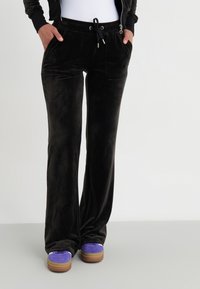 Pantalones amplios, de felpa y negros, con cintura elástica y bolsillos frontales, combinados con zapatillas moradas con suelas marrones.