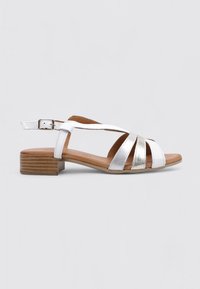 Sandales en cuir blanc avec un petit talon en bois, présentant des lanières entrelacées et un accent métallique argenté. Fermeture à boucle ajustable.