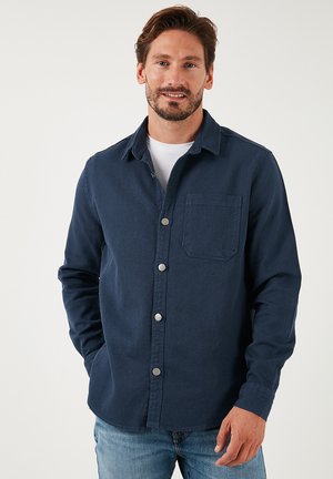Camisa de botones de color azul marino hecha de tela texturizada, con un bolsillo en el pecho y botones plateados. Mangas largas con un diseño de cuello clásico.