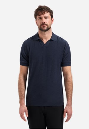 Man met kort krullend haar die een marineblauw poloshirt met korte mouwen en een zwarte broek draagt, staand tegen een effen lichte achtergrond.