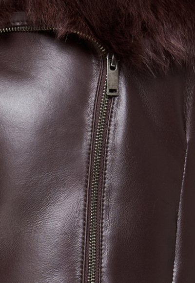 Giubbotto di pelle marrone con colletto in pelliccia, dotato di una zip metallica e una texture liscia. Il design è aderente con un primo piano del dettaglio della zip.