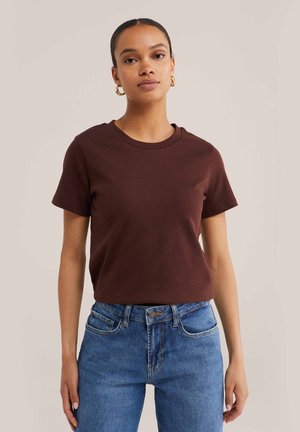 Młoda kobieta o ciemnych włosach, nosząca złote kolczyki koła, brązowy t-shirt z krótkim rękawem i niebieskie jeansy, stojąca na tle jednolitego tła.