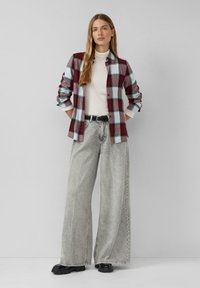 Femme portant une chemise à carreaux rouge et bleu par-dessus un col roulé blanc, un pantalon en jean gris à jambes larges avec une ceinture noire, et des chaussures noires épaisses, debout.