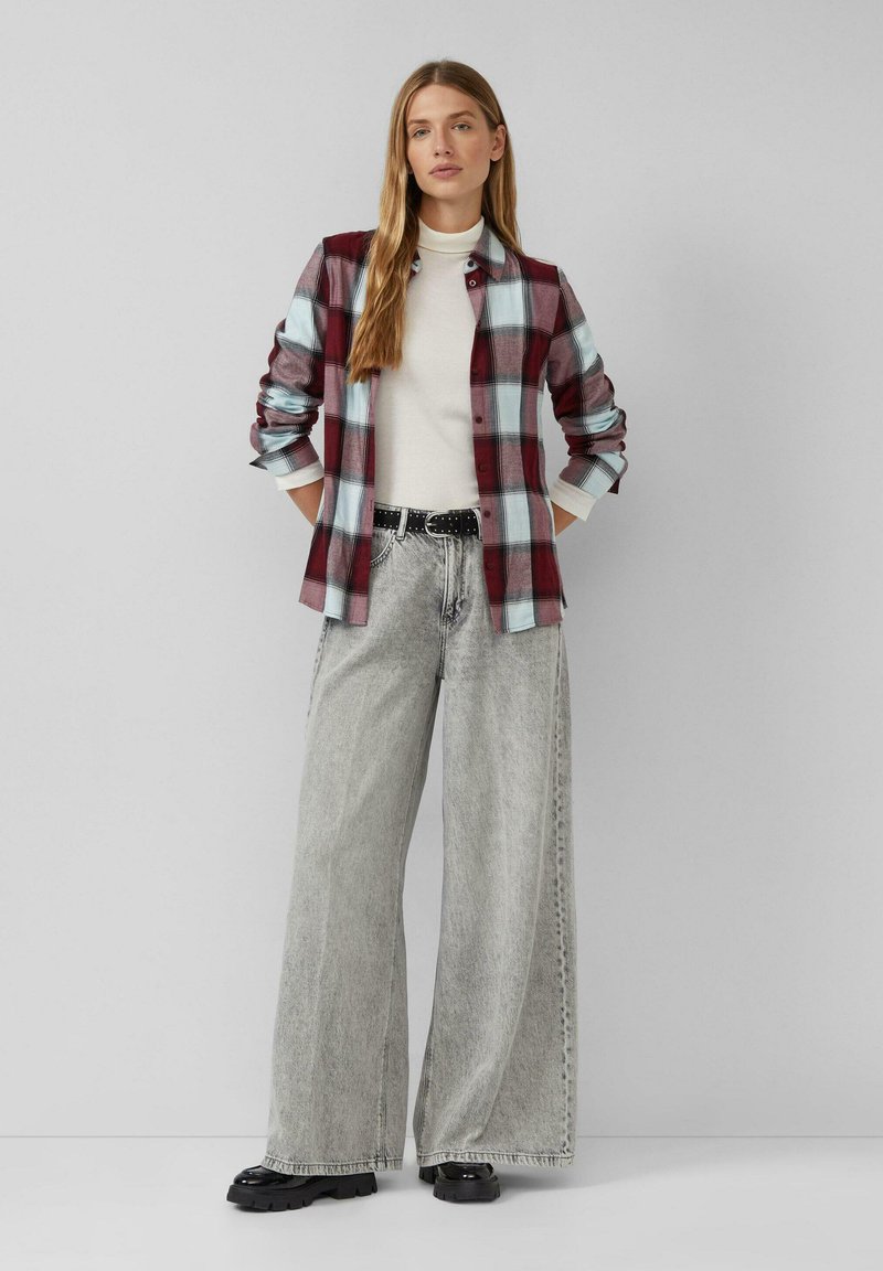 Femme portant une chemise à carreaux rouge et bleu par-dessus un col roulé blanc, un pantalon en jean gris à jambes larges avec une ceinture noire, et des chaussures noires épaisses, debout.