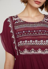 Burgundské šaty s jemnou výšivkou kolem výstřihu a horní části korzetu se květinovými a geometrickými vzory; lehký materiál, krátké rukávy.