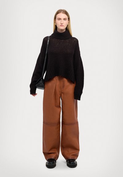 Schwarzer Strickpullover mit hohem Kragen, kombiniert mit lockeren braunen Lederhosen und klobigen schwarzen Schuhen, gehalten mit einer kleinen schwarzen Tasche.