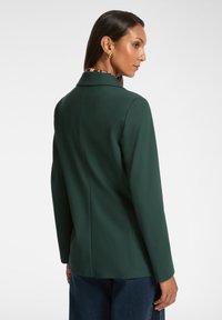 Donna che indossa un blazer verde scuro e jeans blu, vista di spalle e con lo sguardo rivolto sopra la spalla destra su uno sfondo bianco.