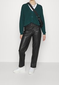 Pintakuvioitu, tummanvihreä neulottu neuletakki sinisillä napeilla, yhdistetty korkeavyötäröisiin mustiin nahkahousuihin ja valkoisiin korollisiin tennareihin.