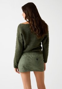 Kobieta z długimi, brązowymi włosami nosi ciemnozielony sweter z dzianiny oraz dopasowaną, zieloną mini spódniczkę, zwrócona plecami w kierunku jednolitego białego tła.