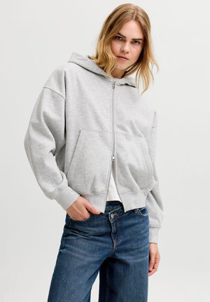 Jeune femme portant un sweat à capuche zippé gris clair et un jean bleu, avec une main dans la poche, debout devant un fond uni.