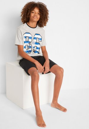 Graues T-Shirt mit blau-weißer Grafik der Nummer "86" und Designelementen, kombiniert mit schwarzen Shorts. Das Modell sitzt barfuß auf einem Würfel.