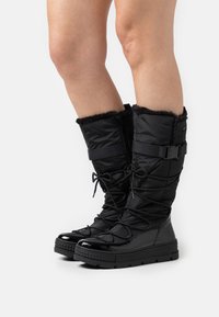 Sorte snø støvler med en puffy tekstiloverdel, pelsfor øverst, justerbar strap og en glanset gummisåle. Har snørelukking.