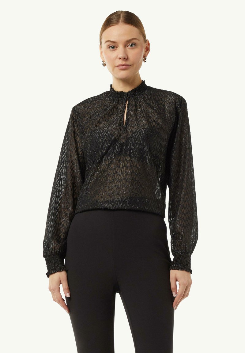 Schwarze Bluse mit einem strukturierten, gemusterten Design, hohem Kragen, Schlüssellochöffnung und elastischen Bündchen, getragen mit schwarzen eng anliegenden Hosen.