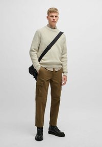 Maglione testurizzato di beige chiaro, pantaloni cargo color khaki, stivali da combattimento neri e una borsa a tracolla nera. Il modello è in piedi contro uno sfondo semplice.
