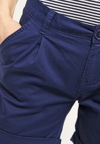 Tmavě modré bavlněné šortky se skladem vpředu, bočními kapsami a zapínáním na knoflík. Tkanina má hladkou strukturu a viditelné švy.