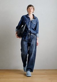 Denim overhemd en losse donkere jeans, gecombineerd met lichtblauwe schoenen. Het model houdt een zwarte, gestructureerde handtas vast tegen een neutrale achtergrond.