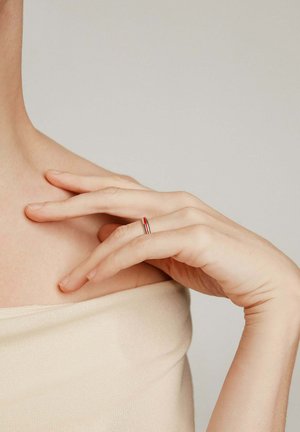 Anillo de plata con acentos rojos y plateados, llevado en una mano delicada que descansa sobre un hombro; fondo de tela neutro.