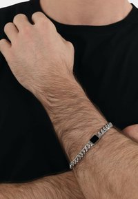 Bracciale in catena d'argento con un accento rettangolare nero, con finitura liscia. Indossato su un polso con peli visibili. Sfondo di una maglietta nera.