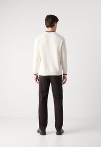 Pull en tricot blanc avec des poignets et un ourlet côtelés, porté avec un pantalon noir et des chaussures noires, vu de l'arrière sur un fond clair.