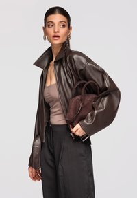 Femme portant une veste en cuir brun foncé et un pantalon noir, tenant un petit sac à main en daim brun foncé, regardant sur le côté contre un fond uni.