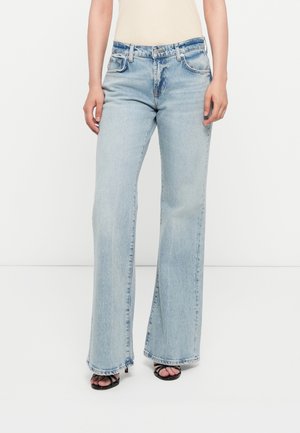 Vrouw draagt lichtblauwe wijd uitlopende jeans en zwarte open-teen hakken, staand met handen ontspannen langs het lichaam op een witte achtergrond.