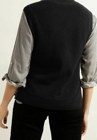 Maglione nero a maglia con un design testurizzato, indossato sopra una camicia a righe bianco e nero, con maniche arrotolate e una finitura liscia.