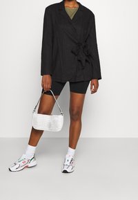 Sort linned blazer med bånd i taljen, båret over en olivengrøn top, parret med sorte shorts og en hvid taske med krokodillemønster. Sølv hardware detaljer.